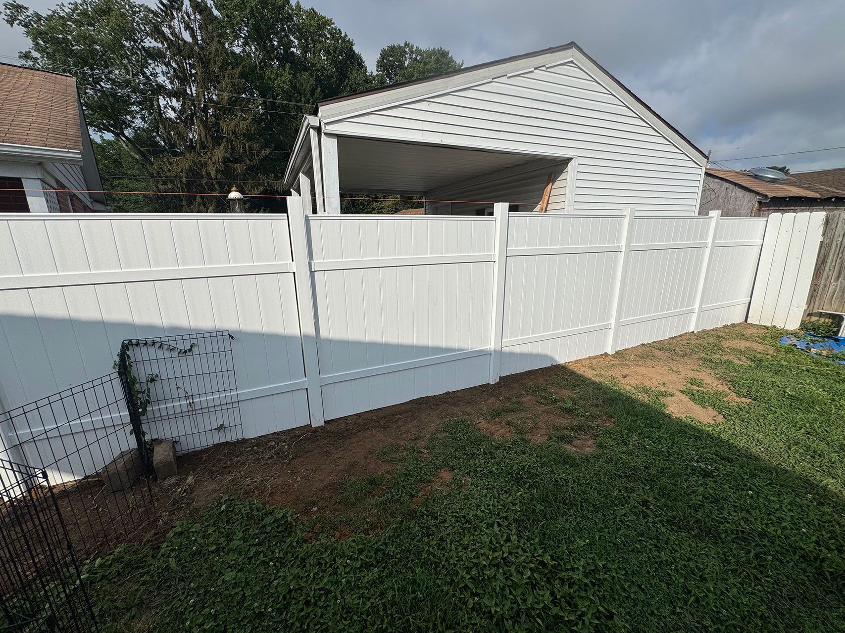 White vinyl fence surrounding a grassy yard with a house and carport in the background under a cloudy sky.
