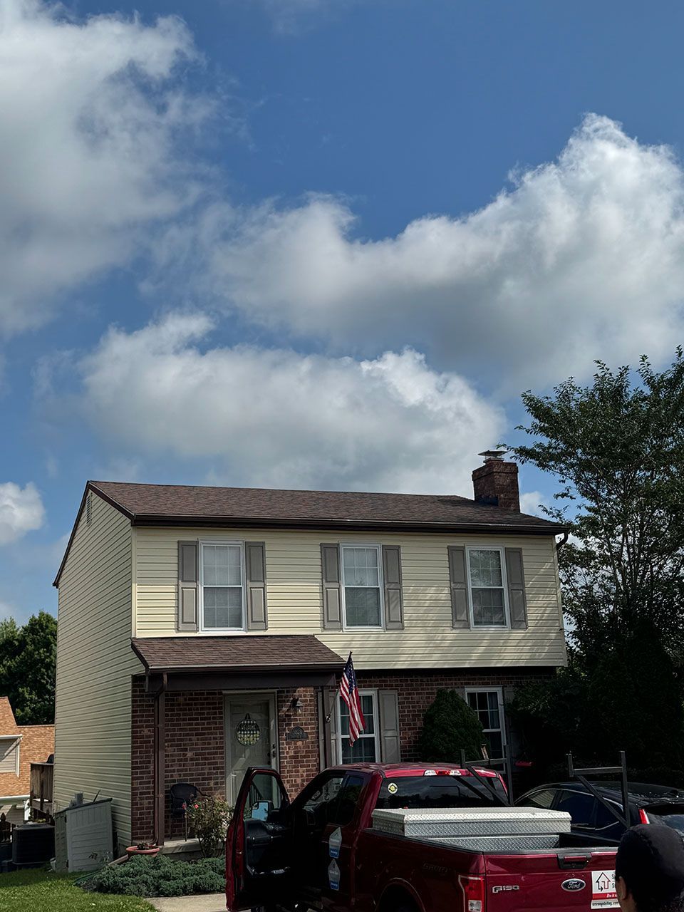 Two-story beige house with brown roof and brick accents; red truck parked in front under a cloudy blue sky.