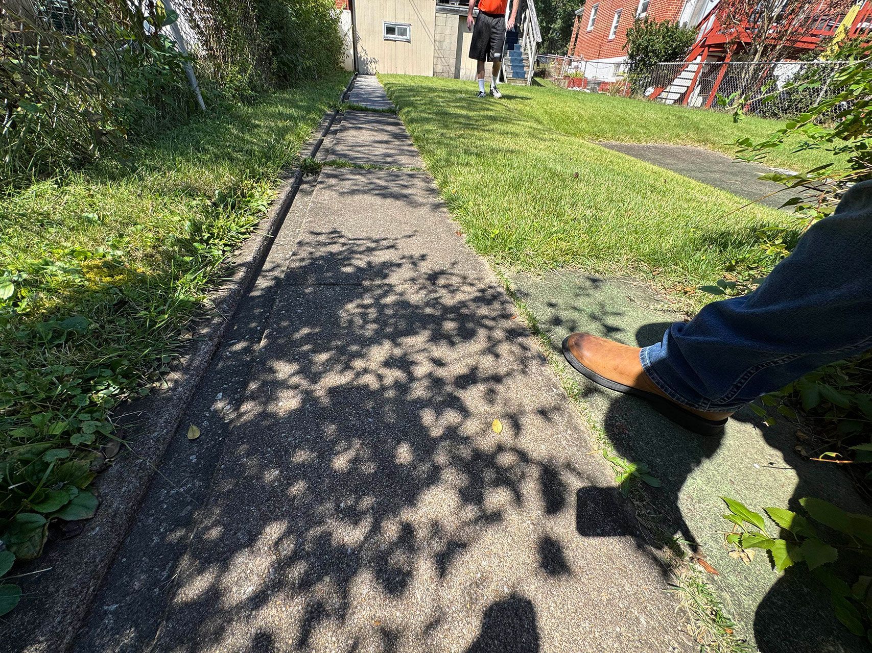 Concrete path through a grassy yard, with a person's leg and shoe in view and a person in the distance.