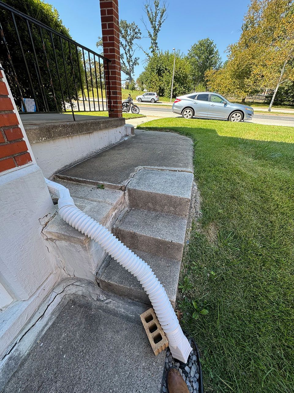 White corrugated pipe extends from a brick home's stoop to the lawn, resting on a brick. Gray steps and a silver car are visible.