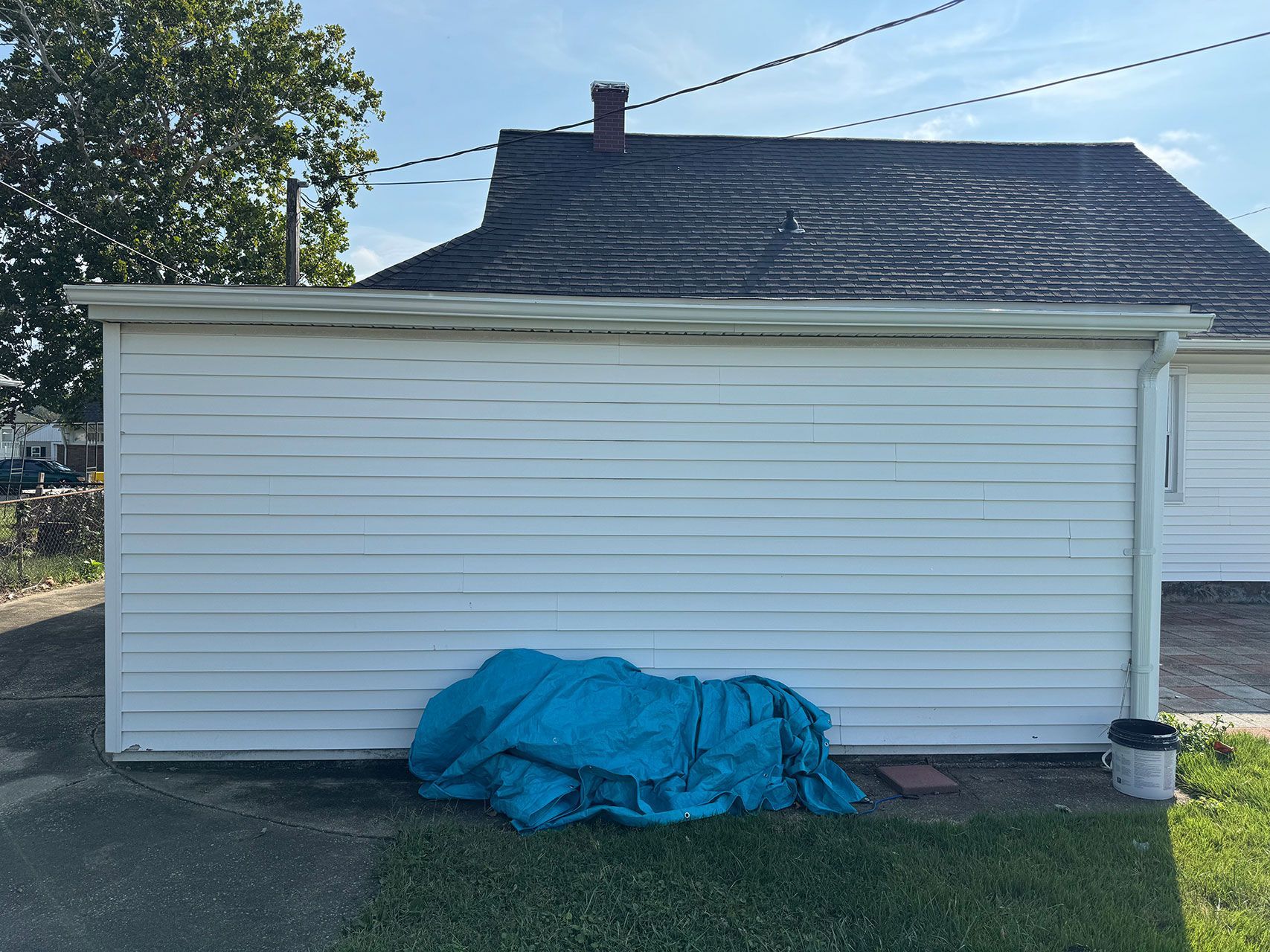 White siding building with a blue tarp in front, next to a house with a dark roof under a blue sky.