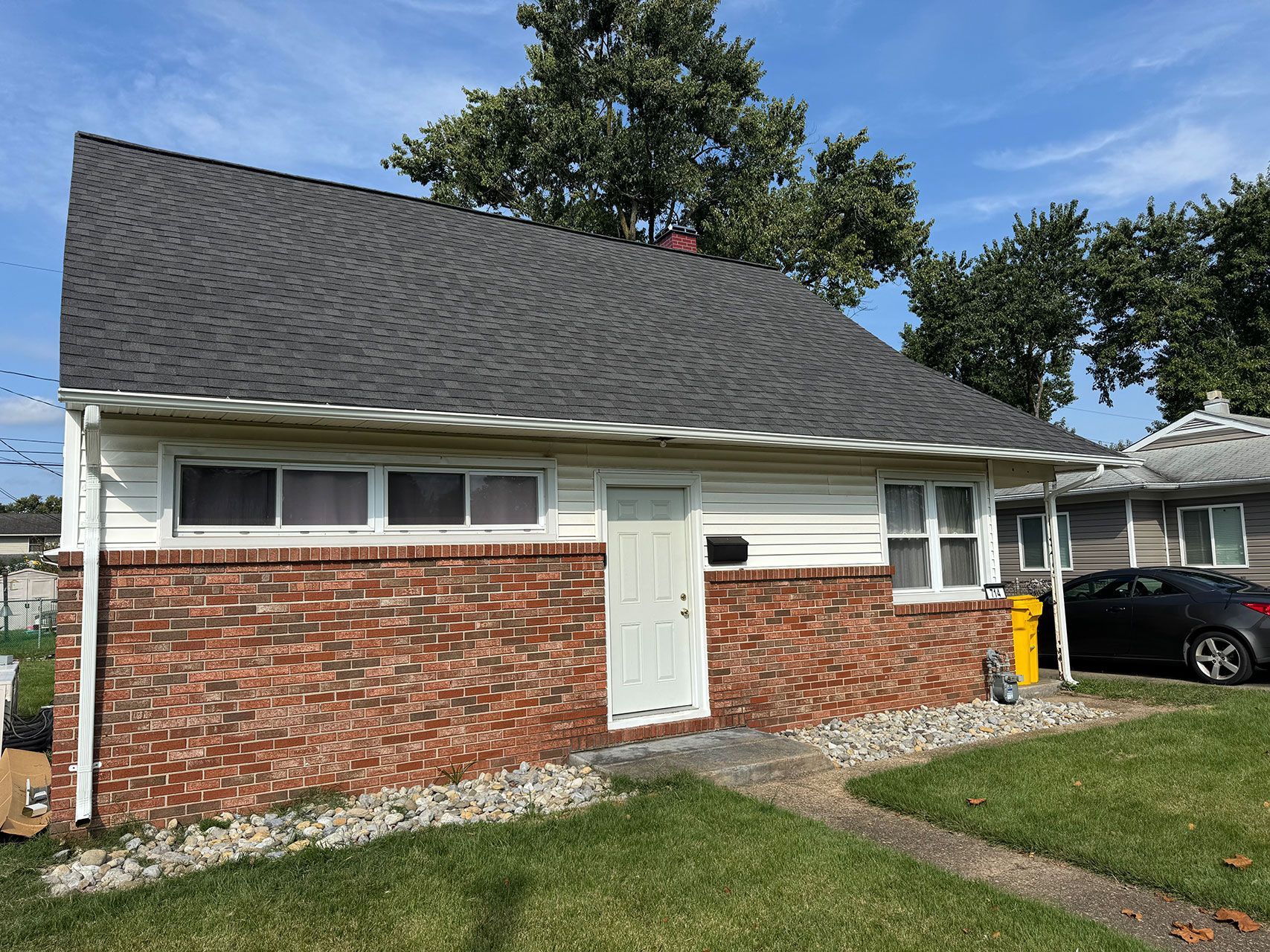 Brick and white-sided house with a dark gray roof. A small lawn and a car are in the setting.