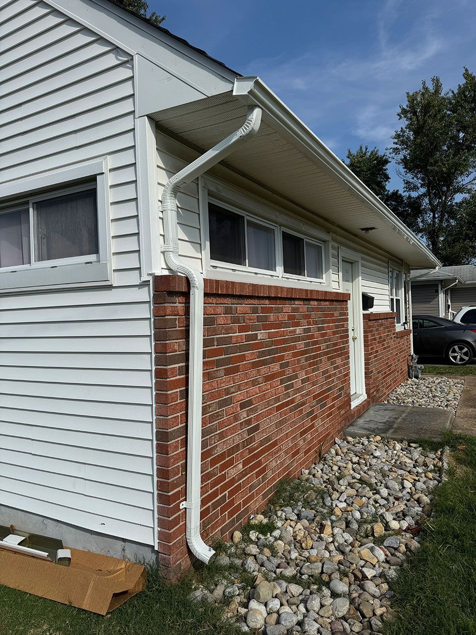 White-sided house with brick wall base and white gutters. Door, windows, and small landscaping with pebbles.