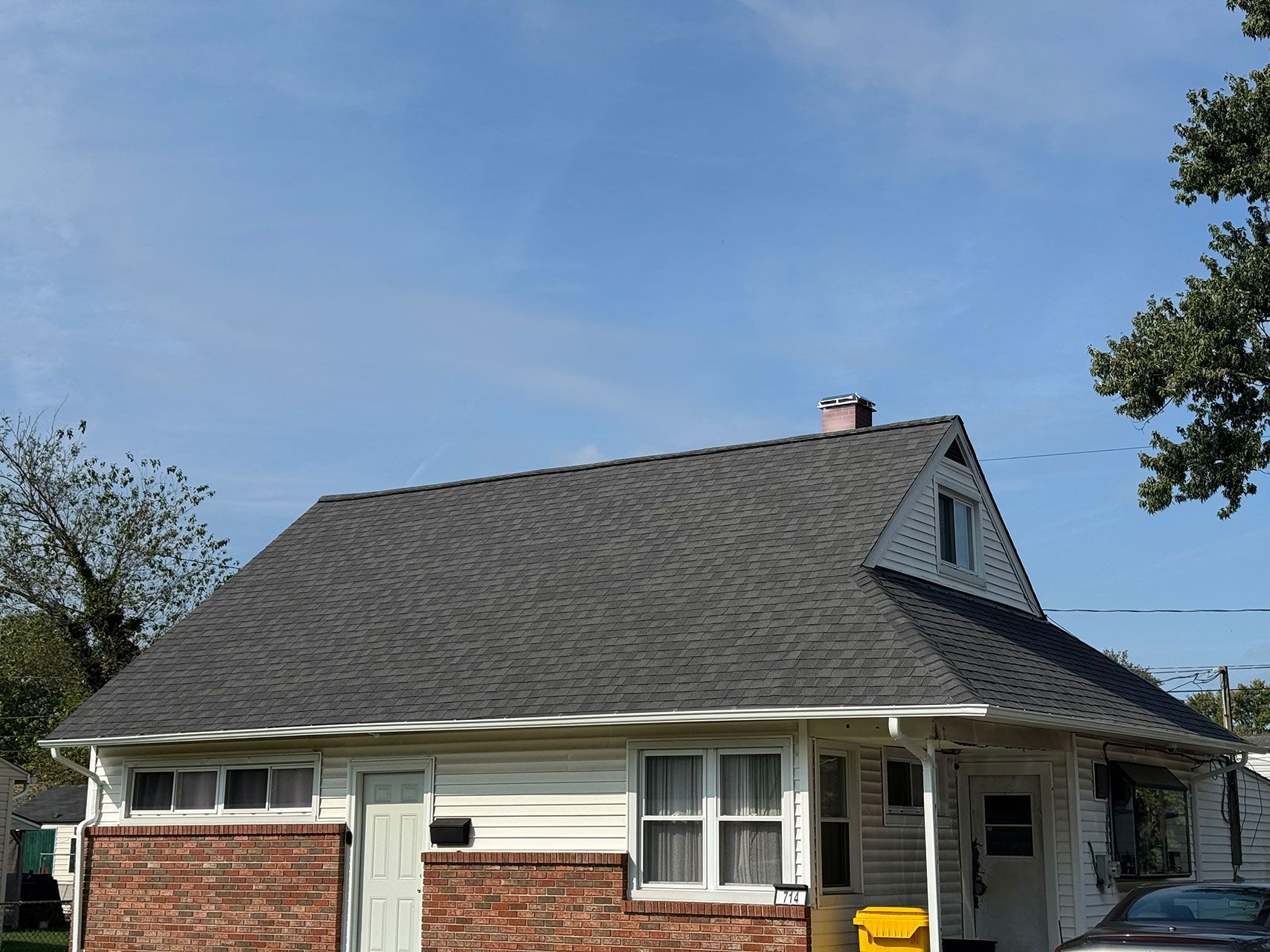 A one-story house with a gray shingled roof, white siding, and brick accents under a blue sky.