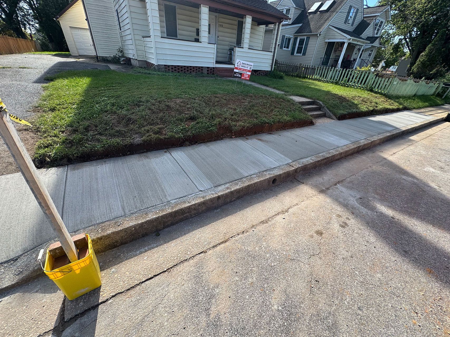 Sidewalk next to a street; a yellow bucket on the left with a wooden pole; houses in the background.