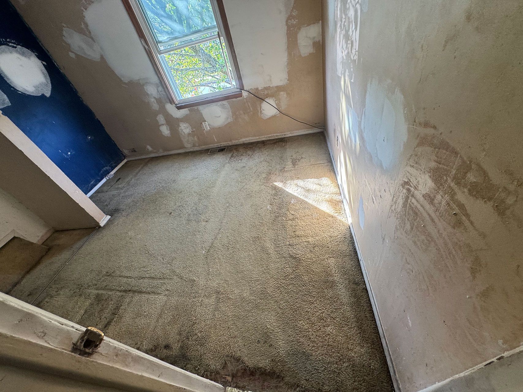 Room with peeling walls, small window, old carpet, and blue door.