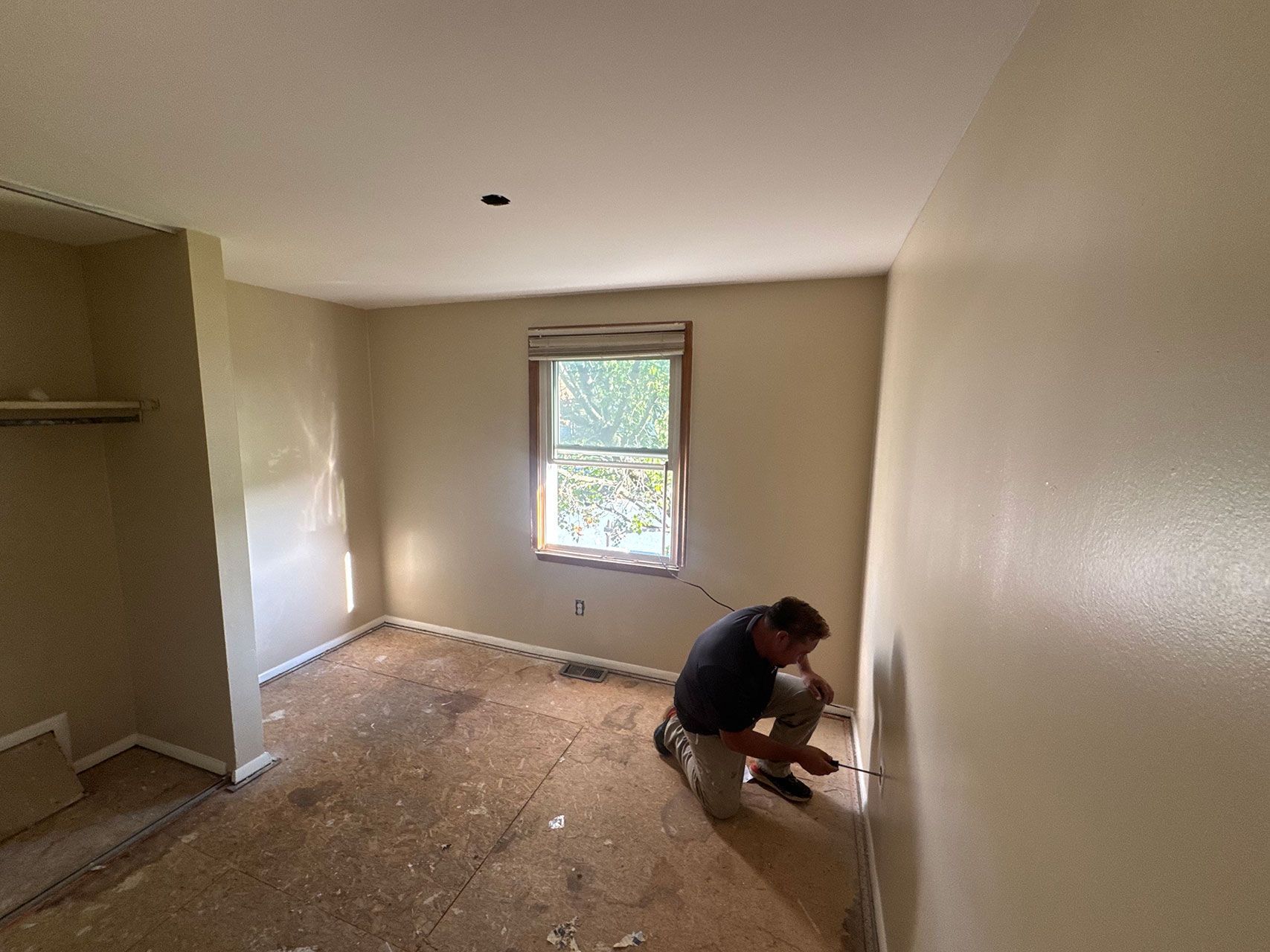 Person kneels to work on the wall in a room with a window, old flooring, and beige walls.