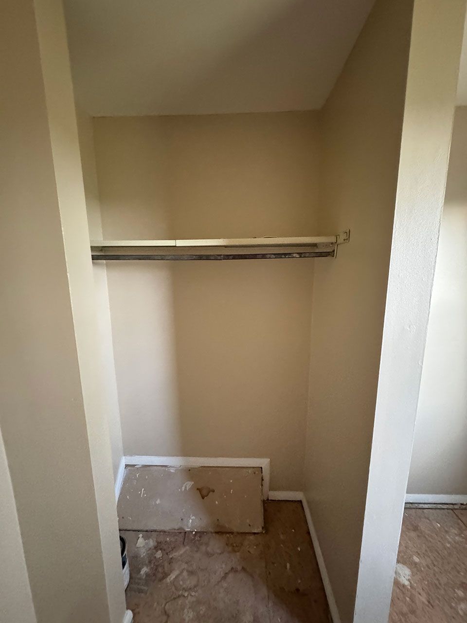 Empty closet with a clothing rod. Beige walls and a damaged floor.