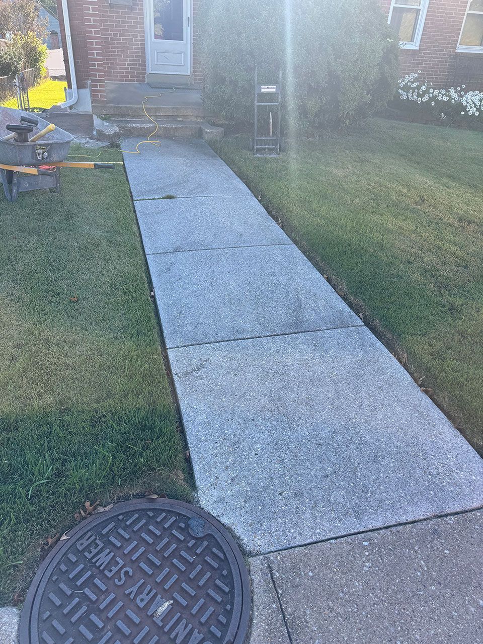 Concrete walkway leading to a house with a manhole cover in the foreground. Green lawn on either side.