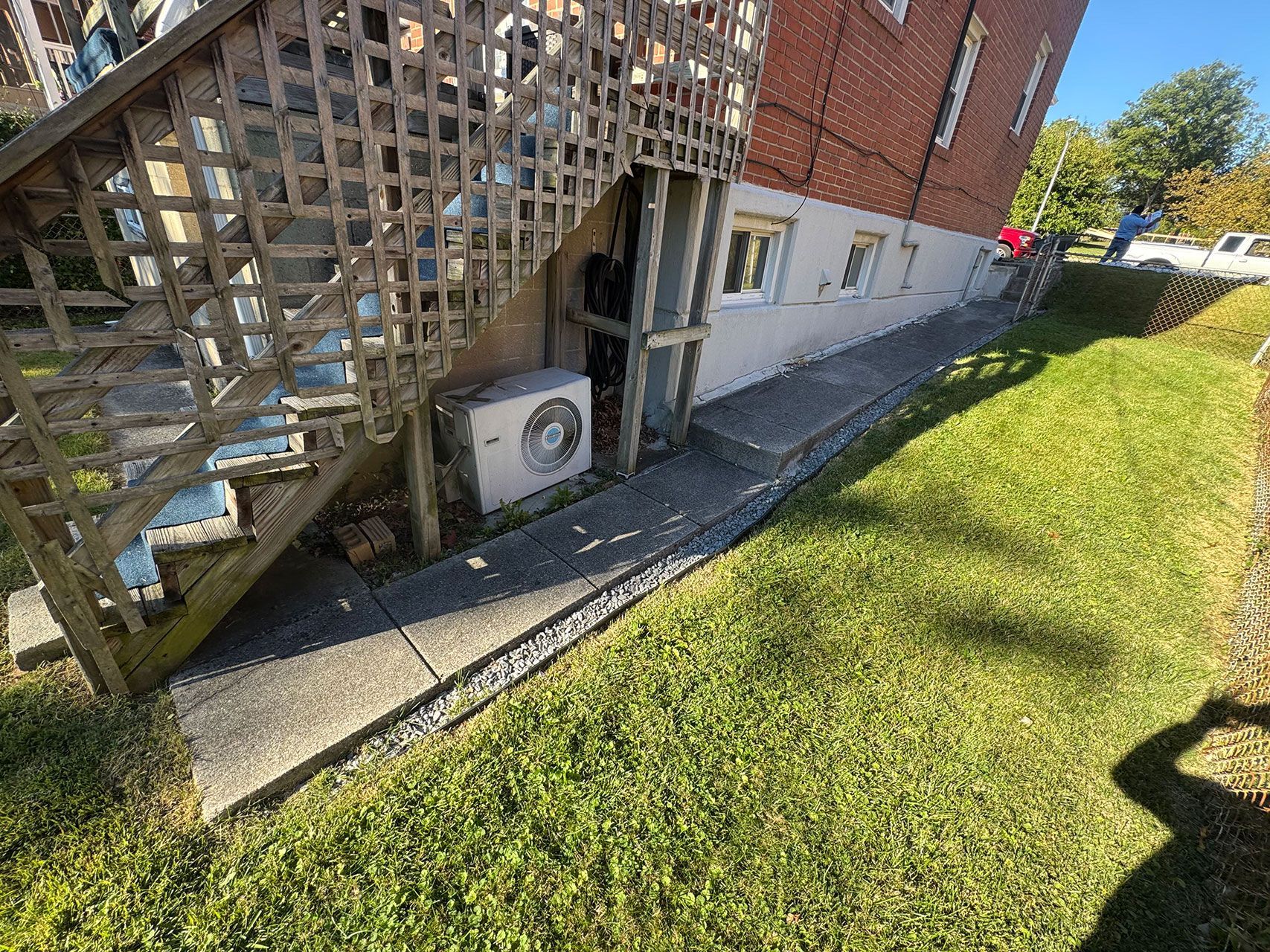 Concrete walkway along a brick building next to a grassy area. Wooden stairs and AC unit.