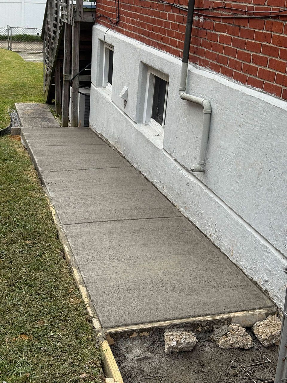 Concrete sidewalk next to a brick building with white stucco base and small windows, leading to stairs.