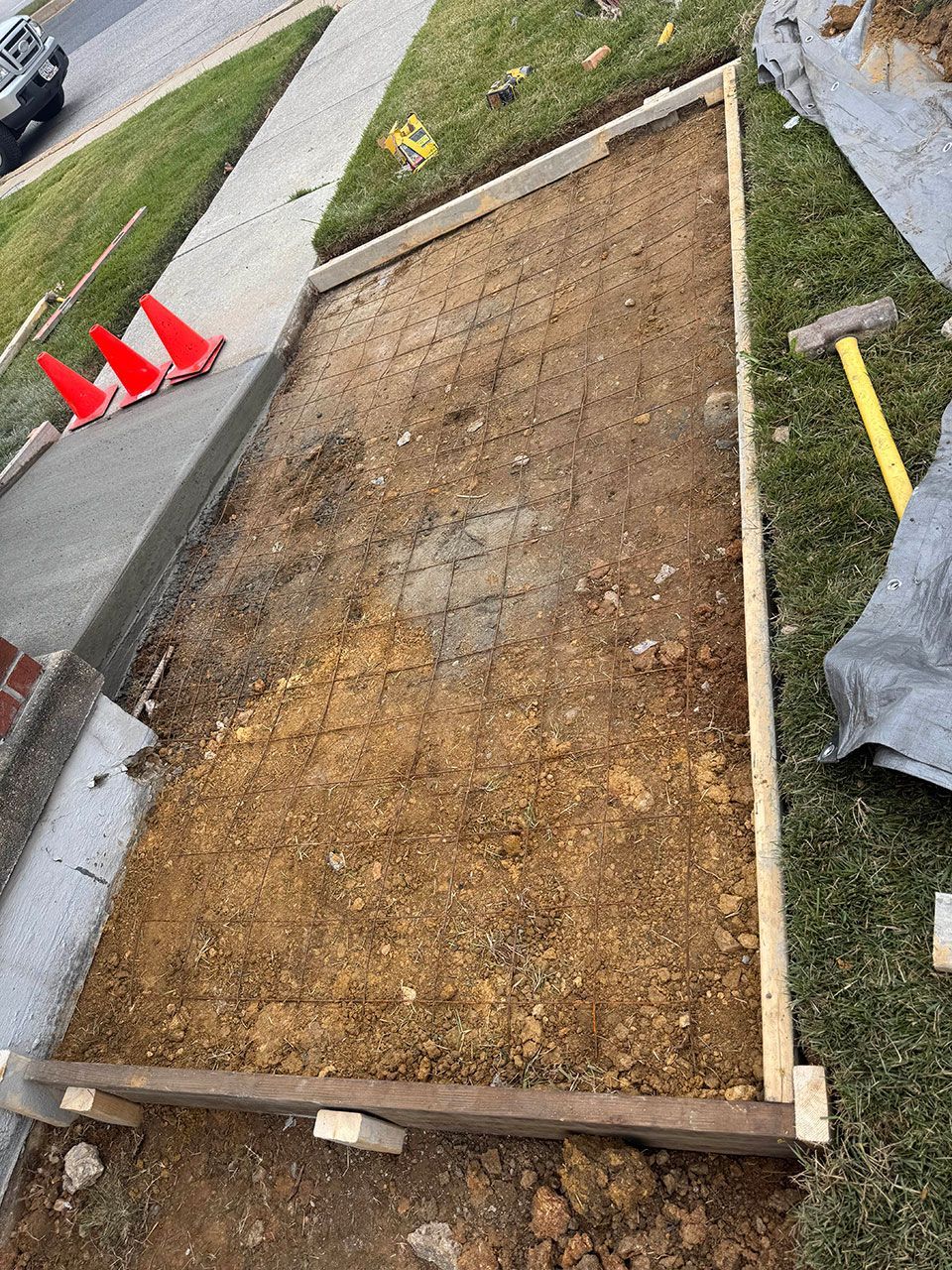 Prepared ground for concrete. Wooden frame and wire mesh in a rectangular dirt patch beside a sidewalk, with orange cones in the background.