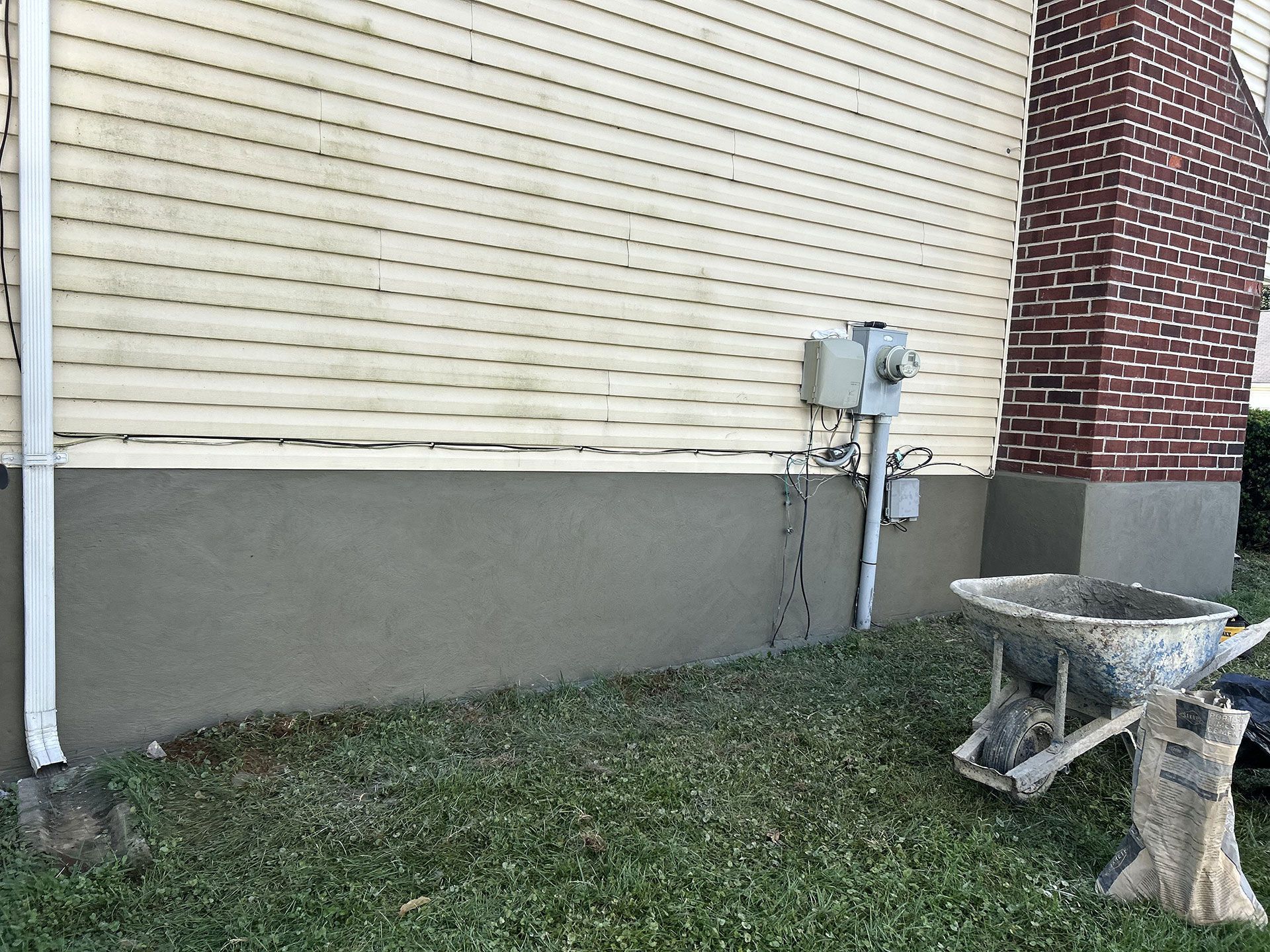 Grey stucco base on a house, below vinyl siding and brick chimney. A wheelbarrow and lawn present.