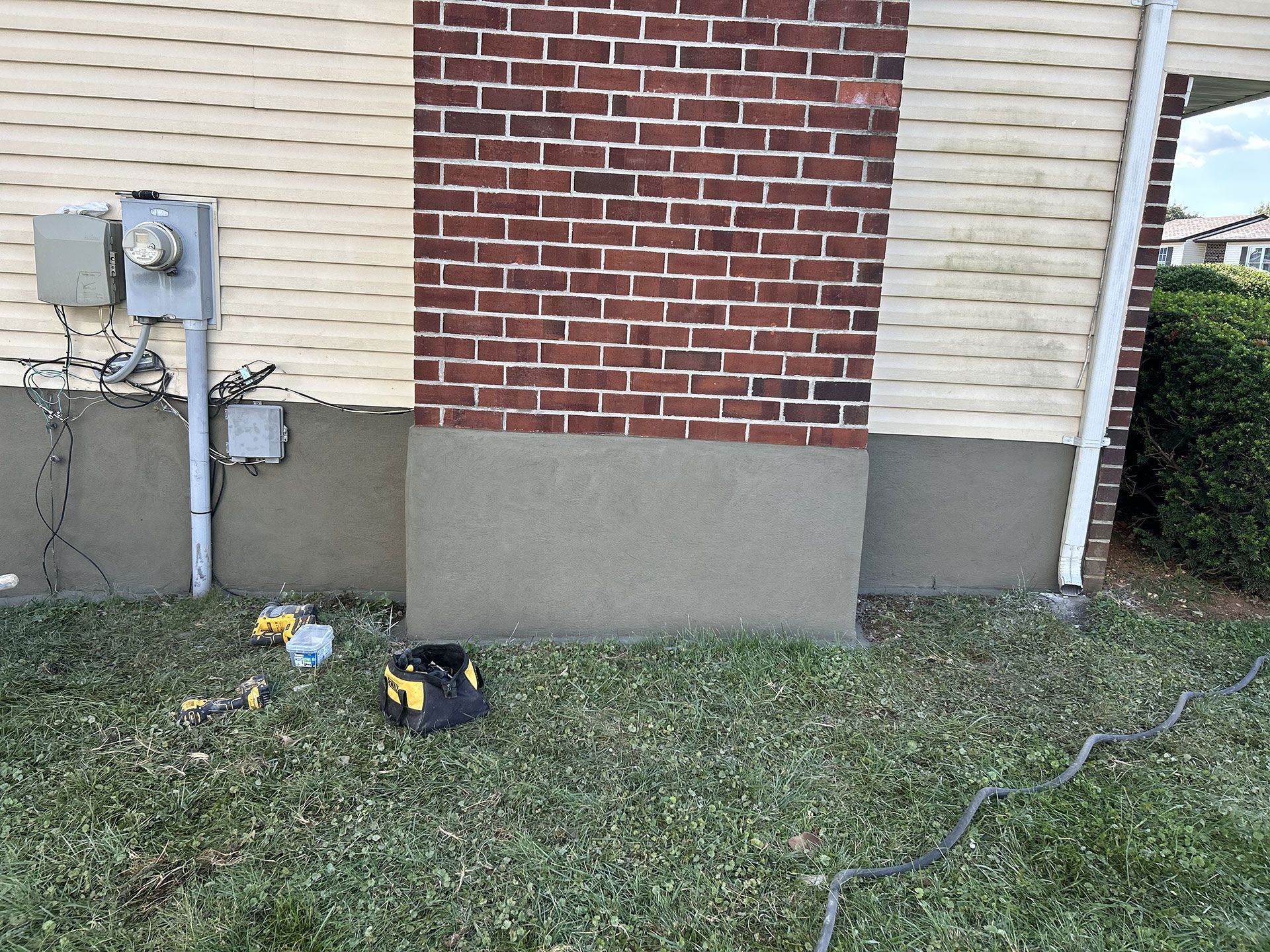 Exterior house wall with brick, siding, and stucco base. Tools on the grass.