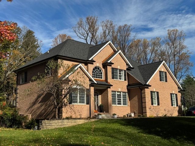 Two-story brick house with a dark roof, green lawn, and trees under a blue sky.