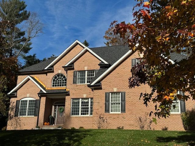 Two-story brick house with a dark roof and black shutters, fall foliage, and a blue sky.