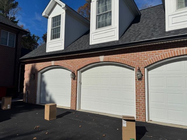 Three white garage doors with brick arches, white trim, and dormers on a dark roof.
