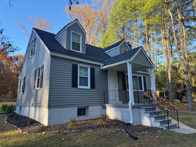 Gray house with black shutters, steps, and porch under a blue sky, surrounded by grass and trees.