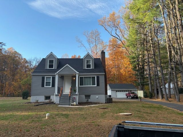 Gray house with black roof and dormers, on a lawn with fall foliage backdrop.