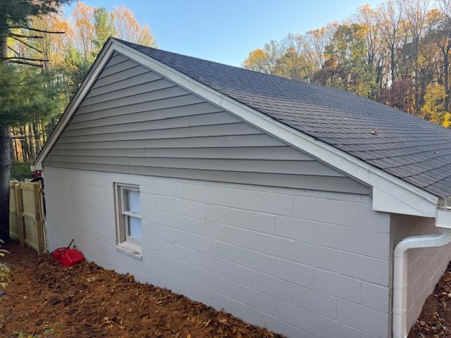 Side view of a building with gray siding, white trim, and a small window. Brown dirt and a wooden fence are below.