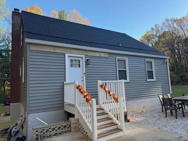 Gray-sided house with a white door, stairs, and a dark roof. Orange decorations hang from the railing.