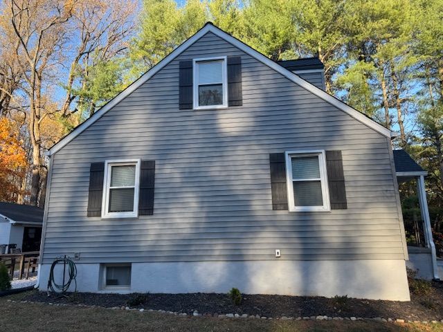 Gray house with white trim, black shutters, and a dark roof; set against a backdrop of trees and a blue sky.