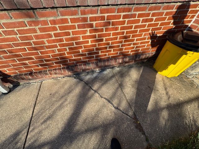 Cracked sidewalk next to a red brick wall and yellow trash can. Sunlight casts long shadows.
