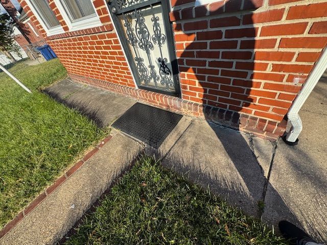 Brick building entrance with a concrete path, a black grate, and shadows cast by sunlight.