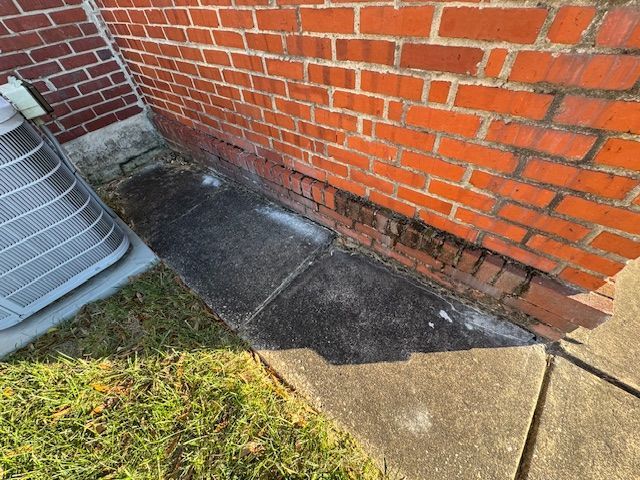 Brick building corner with dark staining at the base, concrete slab, and lawn, next to an AC unit.