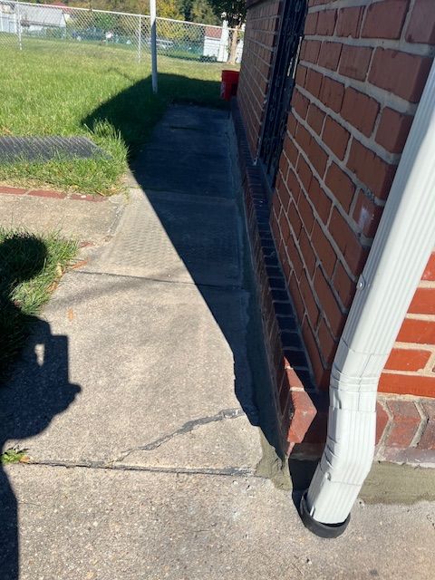 Sidewalk next to a brick building with a white downspout. Shadow of a person present. Green grass and fence in the background.