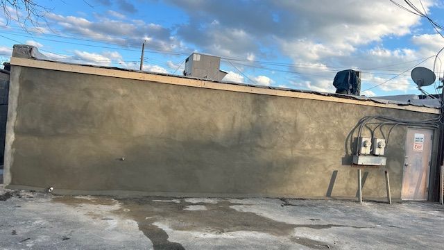 Gray stucco wall with electrical box, water tank, and satellite dish on a rooftop against a cloudy sky.