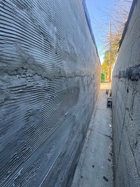Narrow alleyway between two concrete walls, one newly textured with horizontal ridges.