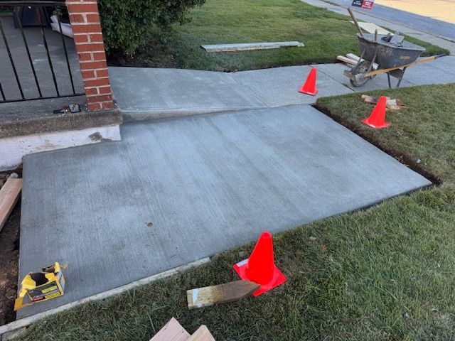 Newly poured concrete sidewalk with orange cones and a wheelbarrow on grass next to a house.