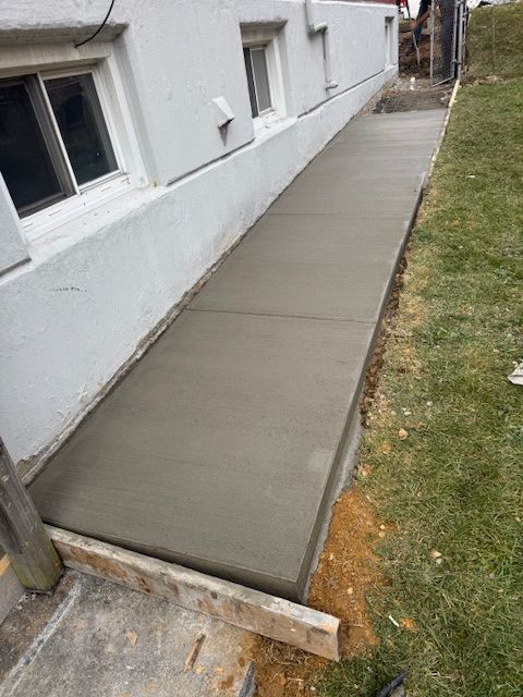 Newly poured concrete sidewalk along a white building foundation, next to a grassy area.