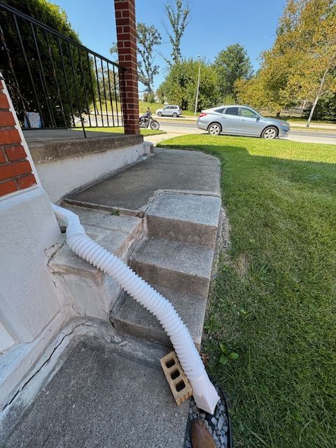 White downspout directs water onto concrete steps, sidewalk, and grass lawn; street with cars in background.