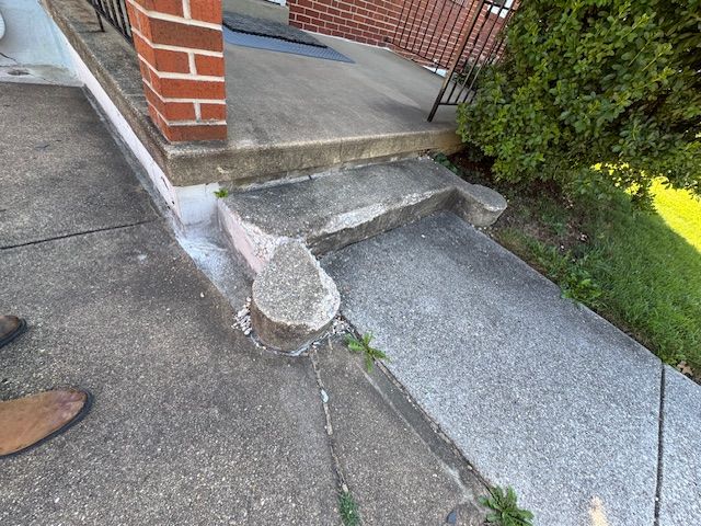 Concrete porch with damaged steps, brick column, and cracked sidewalk. Green bush on right.