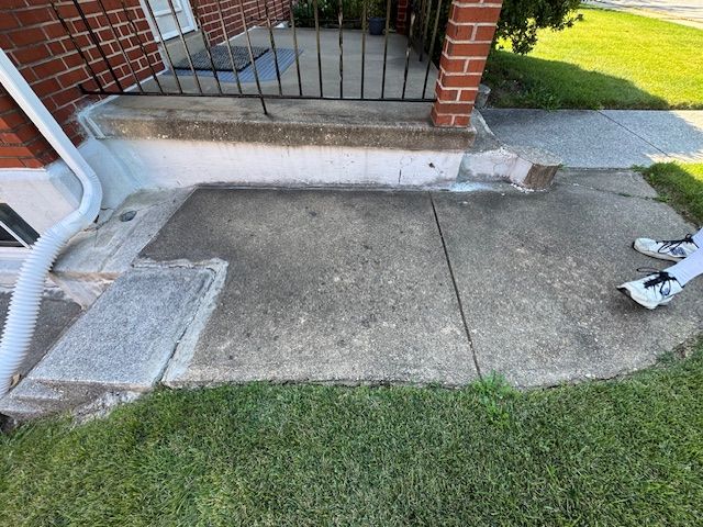 Concrete porch with cracked slab and step near a building with a downspout and wrought iron railing.