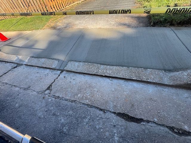 Freshly poured concrete sidewalk next to an older, cracked asphalt surface, with caution tape and an orange cone.