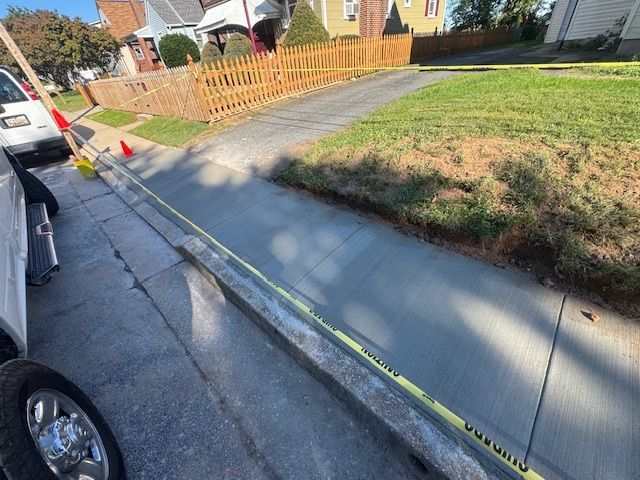 New concrete sidewalk with caution tape, adjacent to a driveway and grassy area, with residential houses in the background.