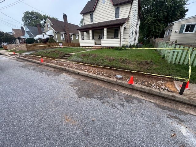 Roadside construction: curb dug up, cones, caution tape, house and lawn in background.
