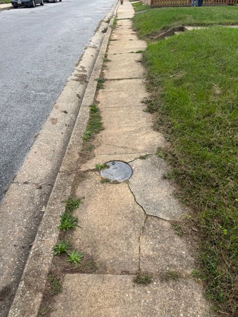 Concrete sidewalk with cracks and weeds, next to a street and grassy lawn.
