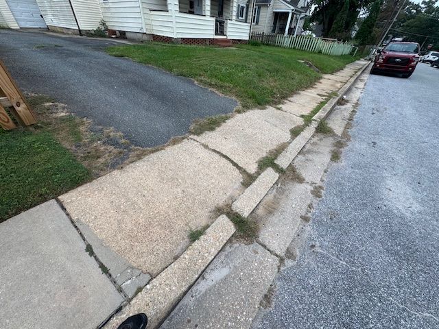 Sidewalk and curb with cracks and grass, along a paved street. A car is parked on the right.