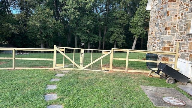 Wooden fence with gate in a yard. Stone house and trees in the background. Green grass and stepping stones.