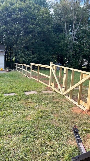 Unfinished wooden fence with gate in grassy backyard, trees in background. Bright, sunny day.