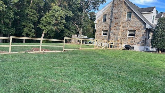 Stone house with new wooden fence in a grassy yard, trees in the background.
