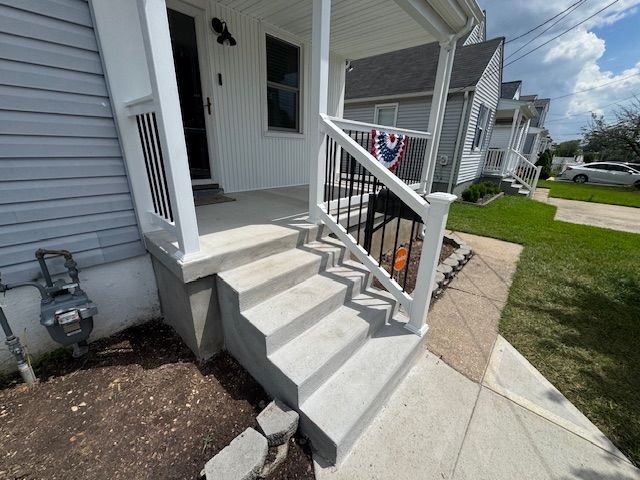 Concrete steps leading to a house's front porch with white railing and black spindles, sidewalk and grass alongside.