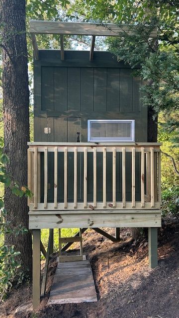 Elevated treehouse with wooden deck and railing. Green walls, white window, steps leading to entrance.