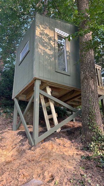 Green treehouse on wooden supports, with windows and a side deck, built on a slope near a tree.