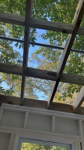 Wooden frame ceiling with open sections. Sky and tree leaves visible. White wall below.
