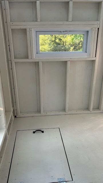 Interior with white walls, a small window, and a floor hatch. Framing is visible.
